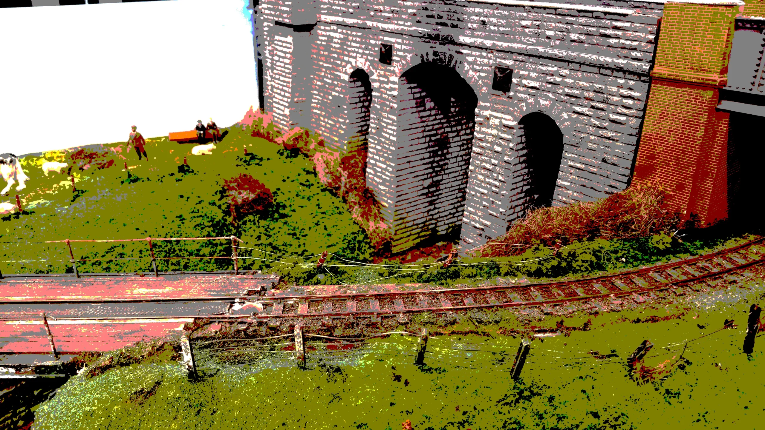 High-contrast model railway scene with a stone viaduct, curving tracks, and miniature figures on a grassy bank